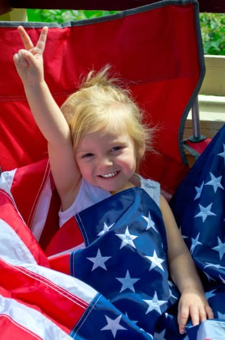 Little girl in chair giving peace sign draped in American Flag